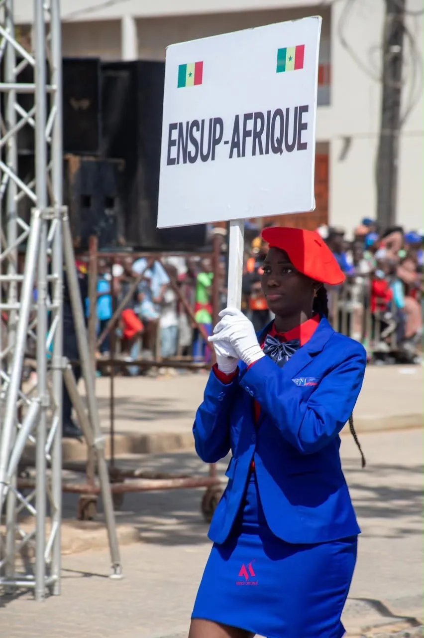 Une étudiante d'ENSUP Afrique en uniforme bleu, béret rouge et gants blancs porte la pancarte « ENSUP-AFRIQUE » lors d'un défilé.